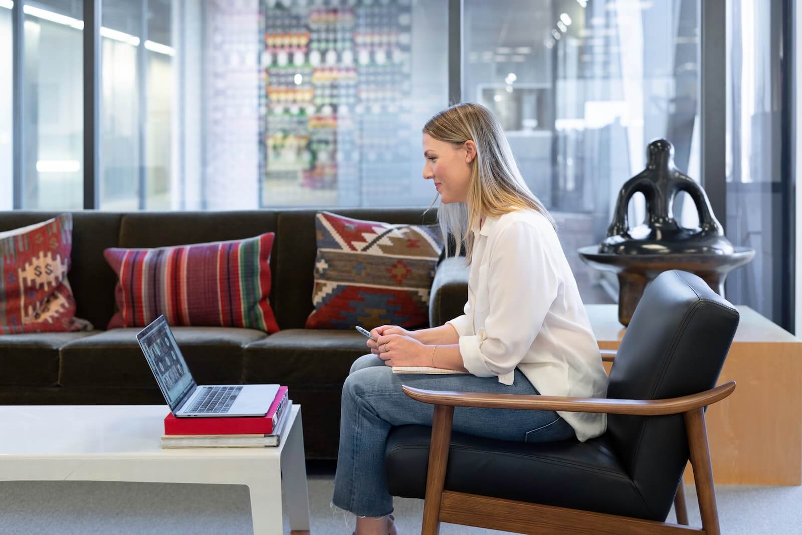 Smiling blonde woman in white shirt and blue denim jeans sitting on a black leather chair looking at a laptop on a coffee table