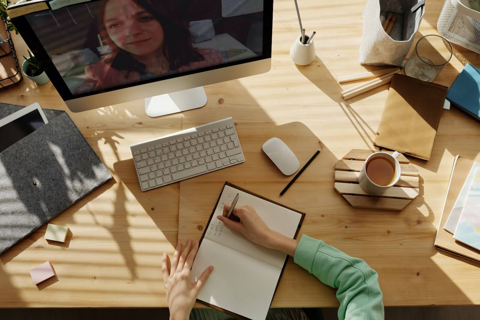 Overhead shot of woman writing in a notebook on a wooden desk while on a video call on a desktop screen