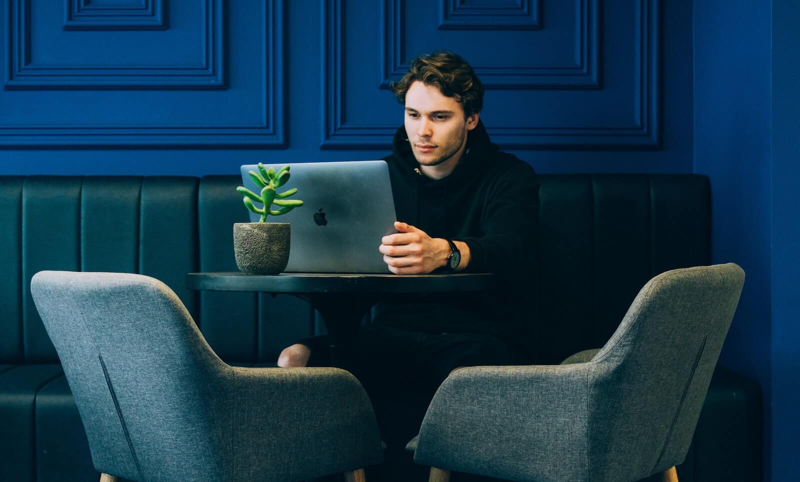 Young man using laptop sitting on small circular table against petrol blue wall
