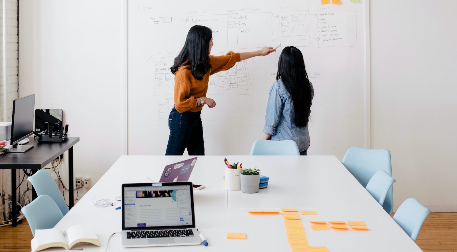 Two women using a write-on wall to collaborate in an office meeting room in front of a meeting table covered with sticky notes