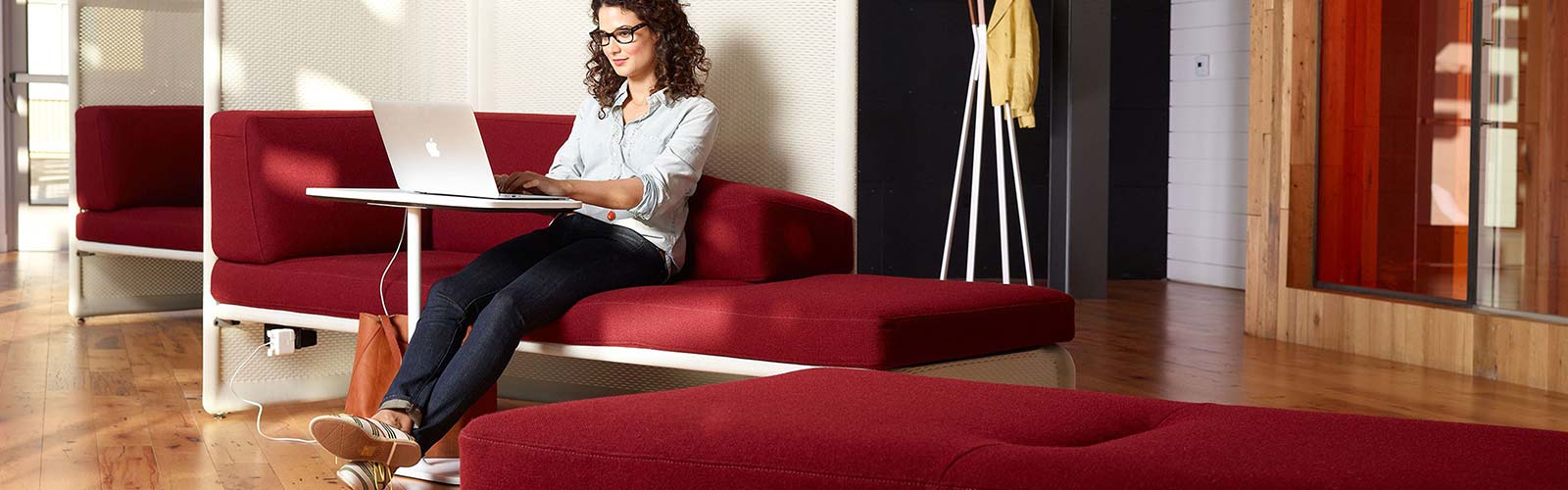 Woman with curly hair in dark trousers and light shirt working on a laptop on a red chair