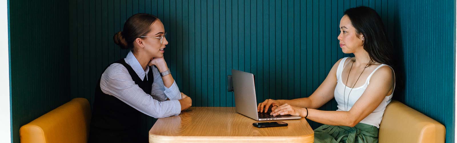 Two female workers sitting across from each other at a table, one with a laptop and the other talking to her