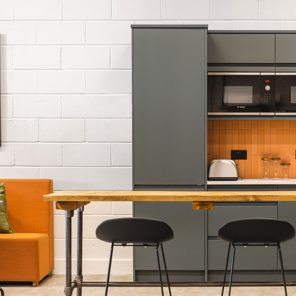 Modern office kitchen design with grey units, orange tiles, bar stools and orange sofa