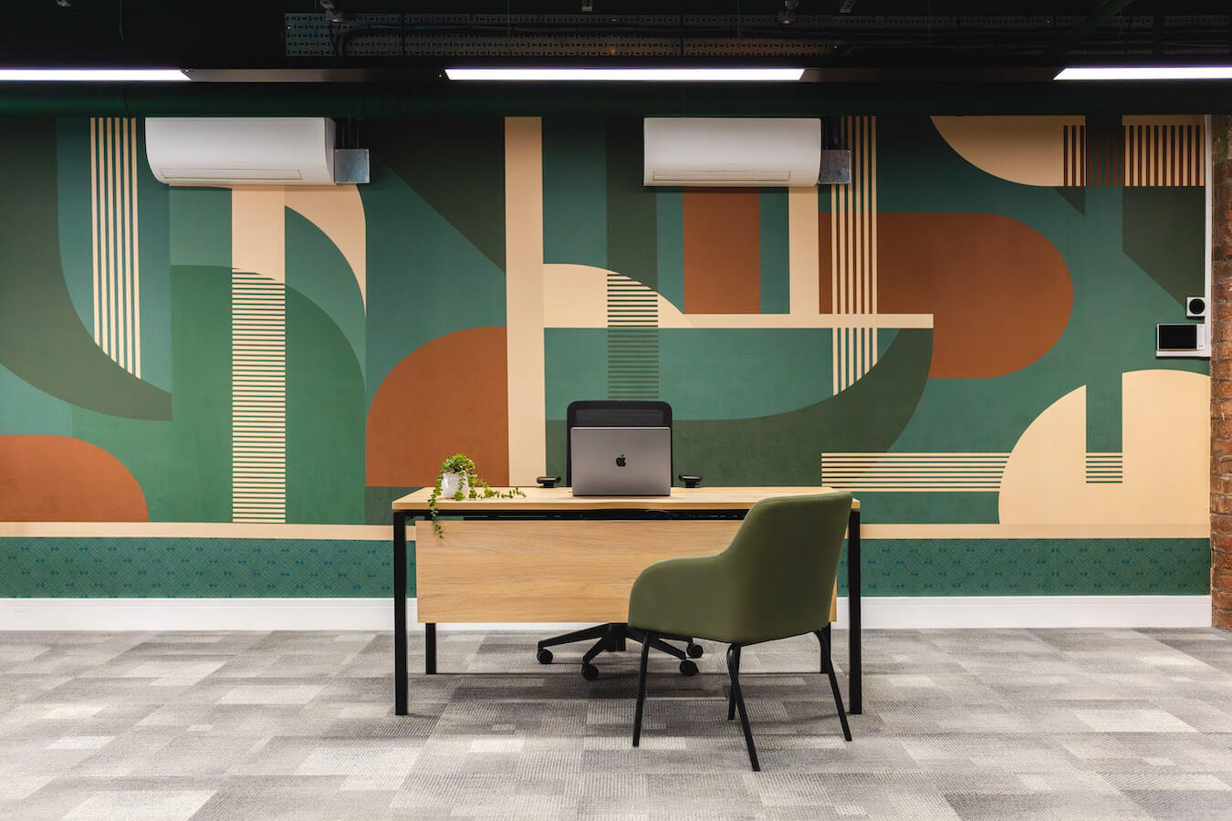 Light wooden desk and green office chair in front of a colourful geometric wallpaper and grey carpet tiles