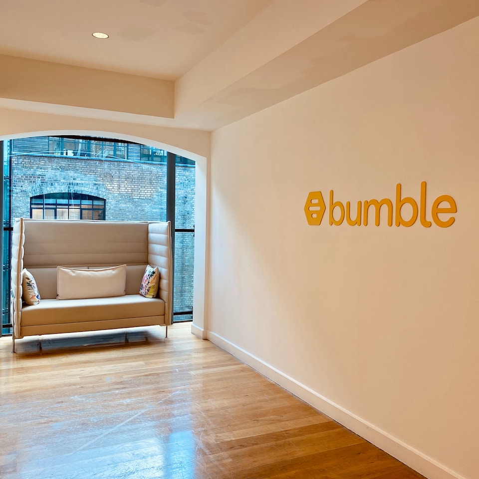 White wall with Bumble logo on it with a floor to ceiling window and grey soft seat