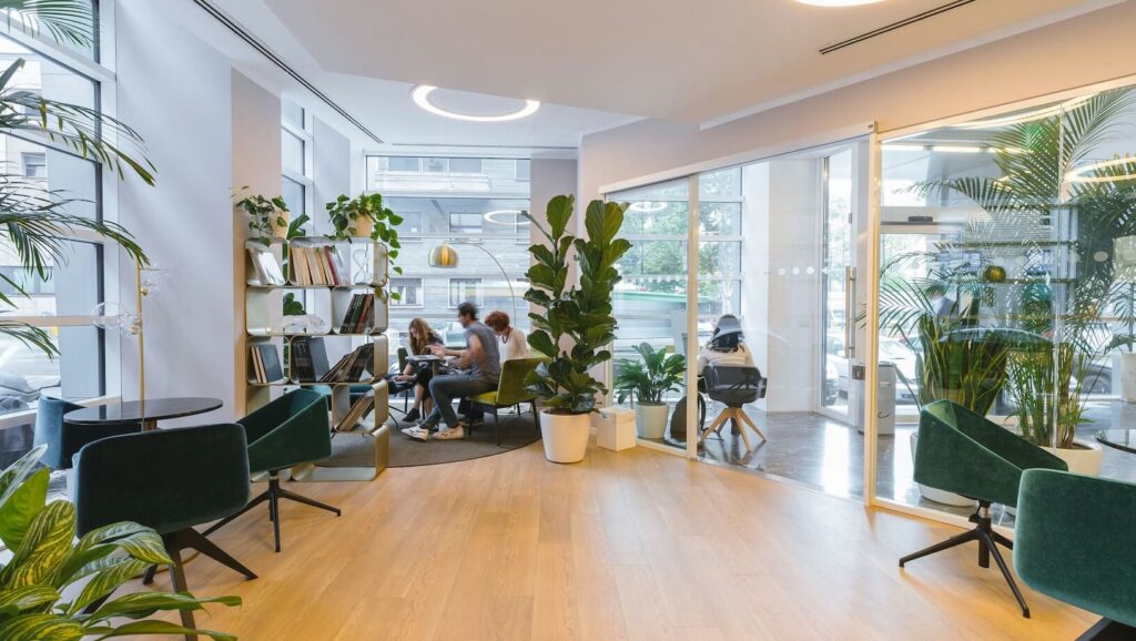 Modern office with light wood floor, glass walls and green indoor plants and people sitting around table