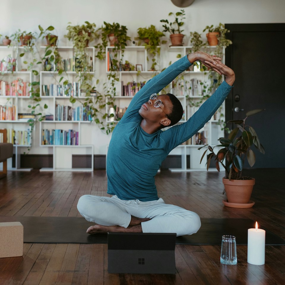 Young man doing a yoga stretch sat on a black yoga mat on a wooden floor with a living wall behind him