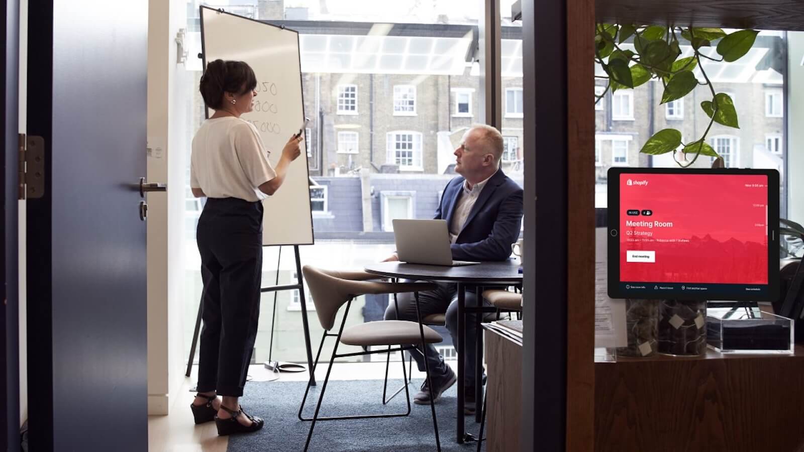 A woman standing next to a flipchart, in front of a man in a blue suit sat at a desk. To the right, is a room booking system mounted to the glass meeting room wall. 